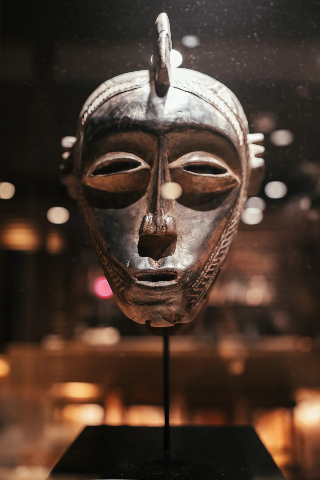 Se ve una máscara africana de metal sobre un soporte, con luces desenfocadas al fondo.