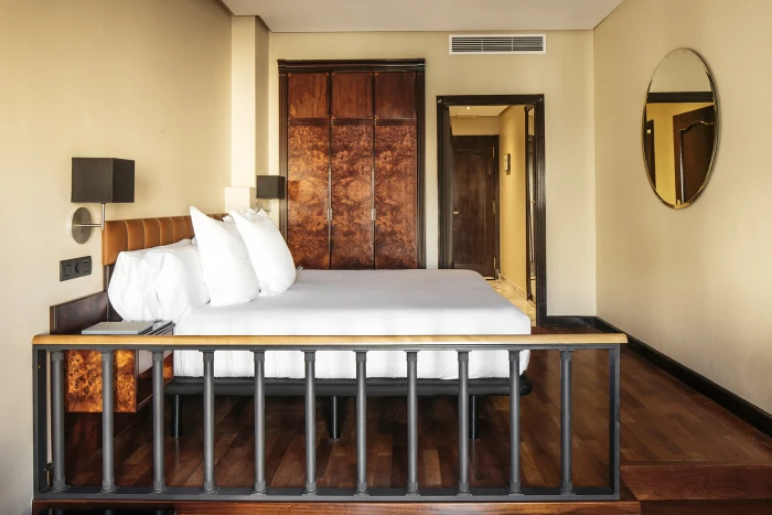 Se ve una cama doble con cabecero de madera, mesita y lámpara, junto a una pared con puerta y un espejo redondo.