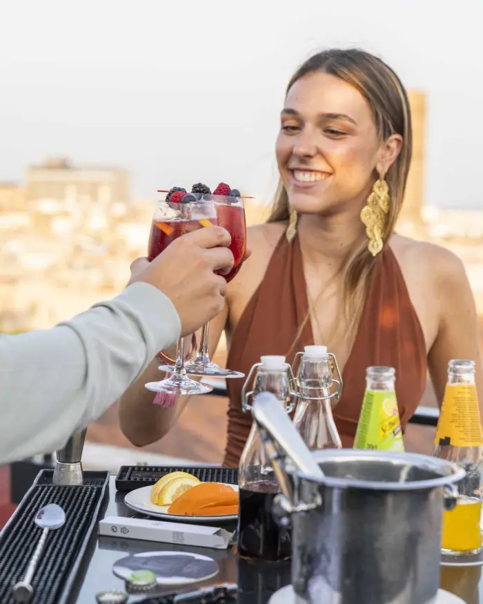 Si vedono mani che preparano bevande con uno shaker e dei bicchieri su un tavolo in una terrazza all'aperto.