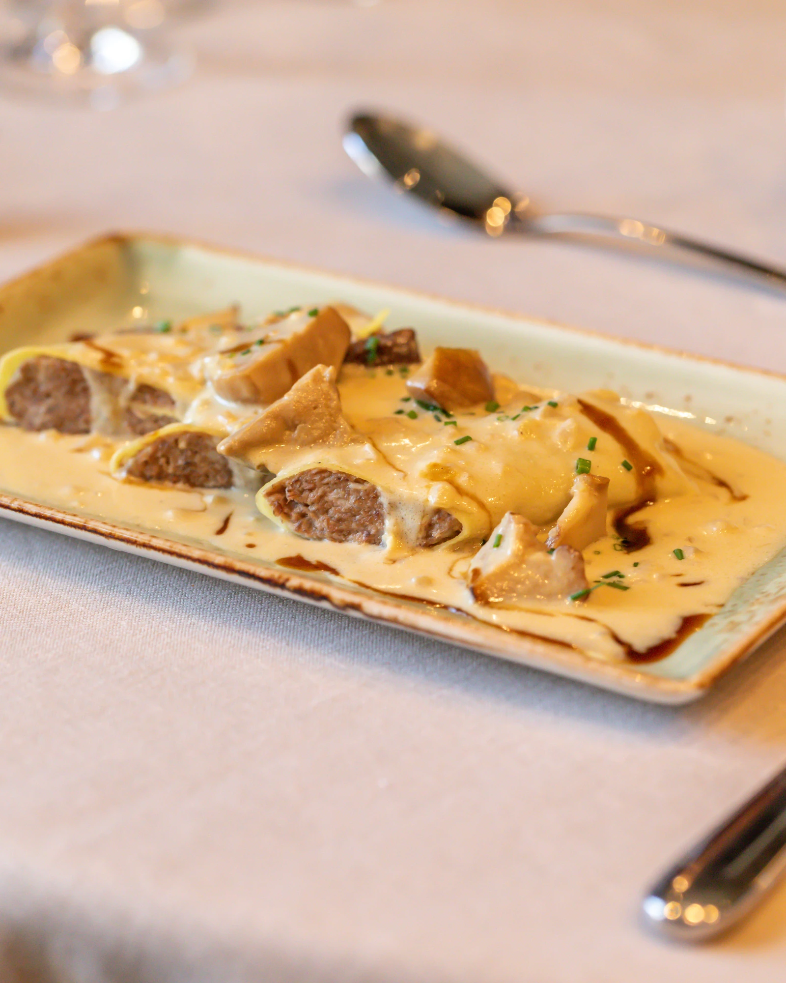 Auf einem rechteckigen Teller liegt eine goldbraune Crêpe mit Käse und Pilzstückchen, garniert mit gehackten Kräutern.
