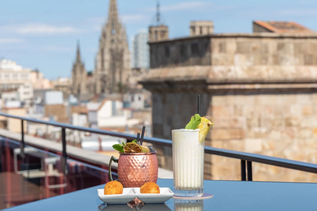 On voit une table avec un plateau de fruits et des verres posés sur une rambarde, sur une terrasse avec des bâtiments à l’arrière-plan.