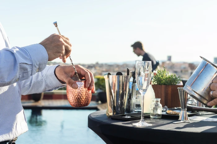 Hands are seen preparing drinks with a shaker and glasses on a table on an outdoor terrace.