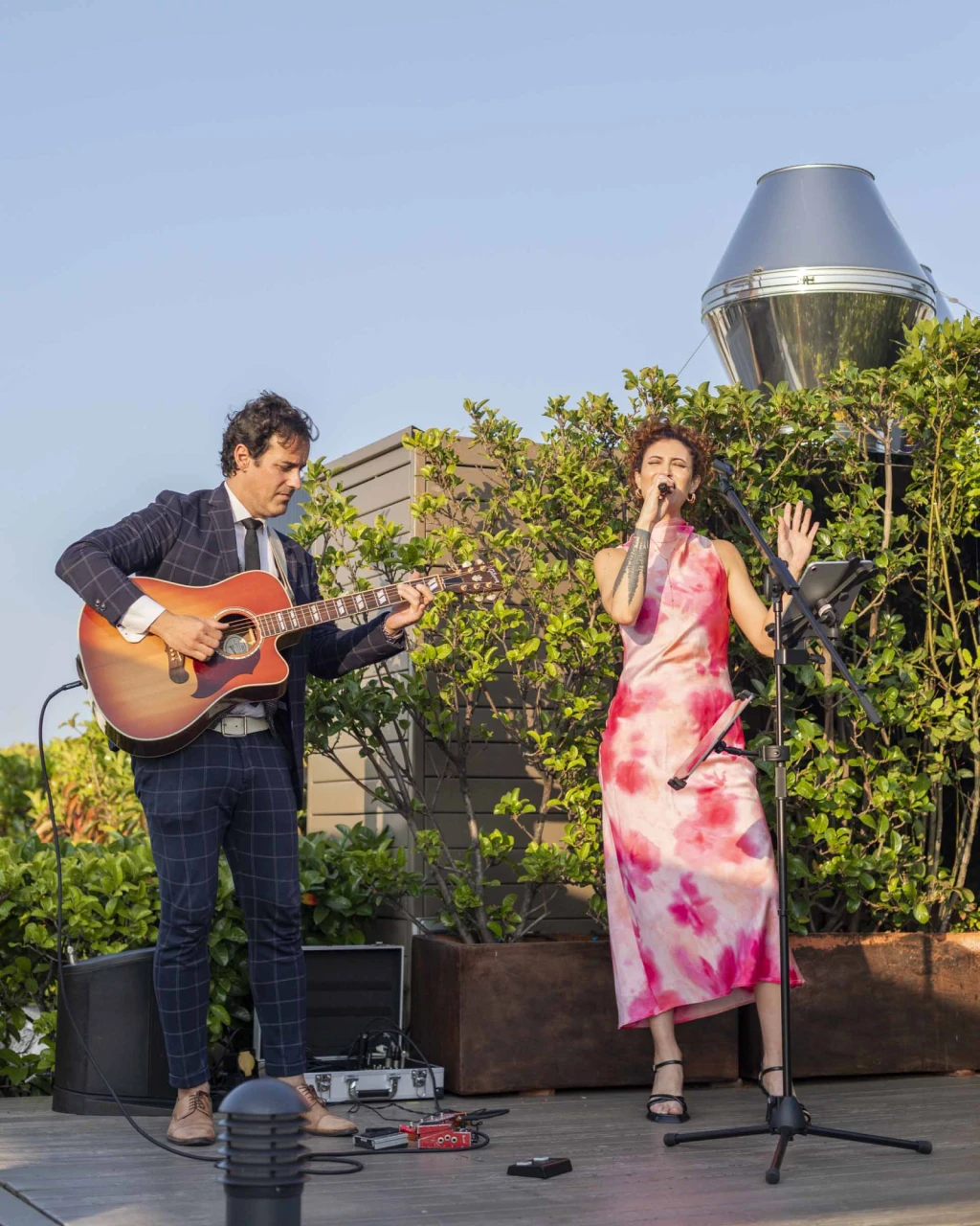 On an outdoor stage, two musicians perform with a singer and an acoustic guitar.