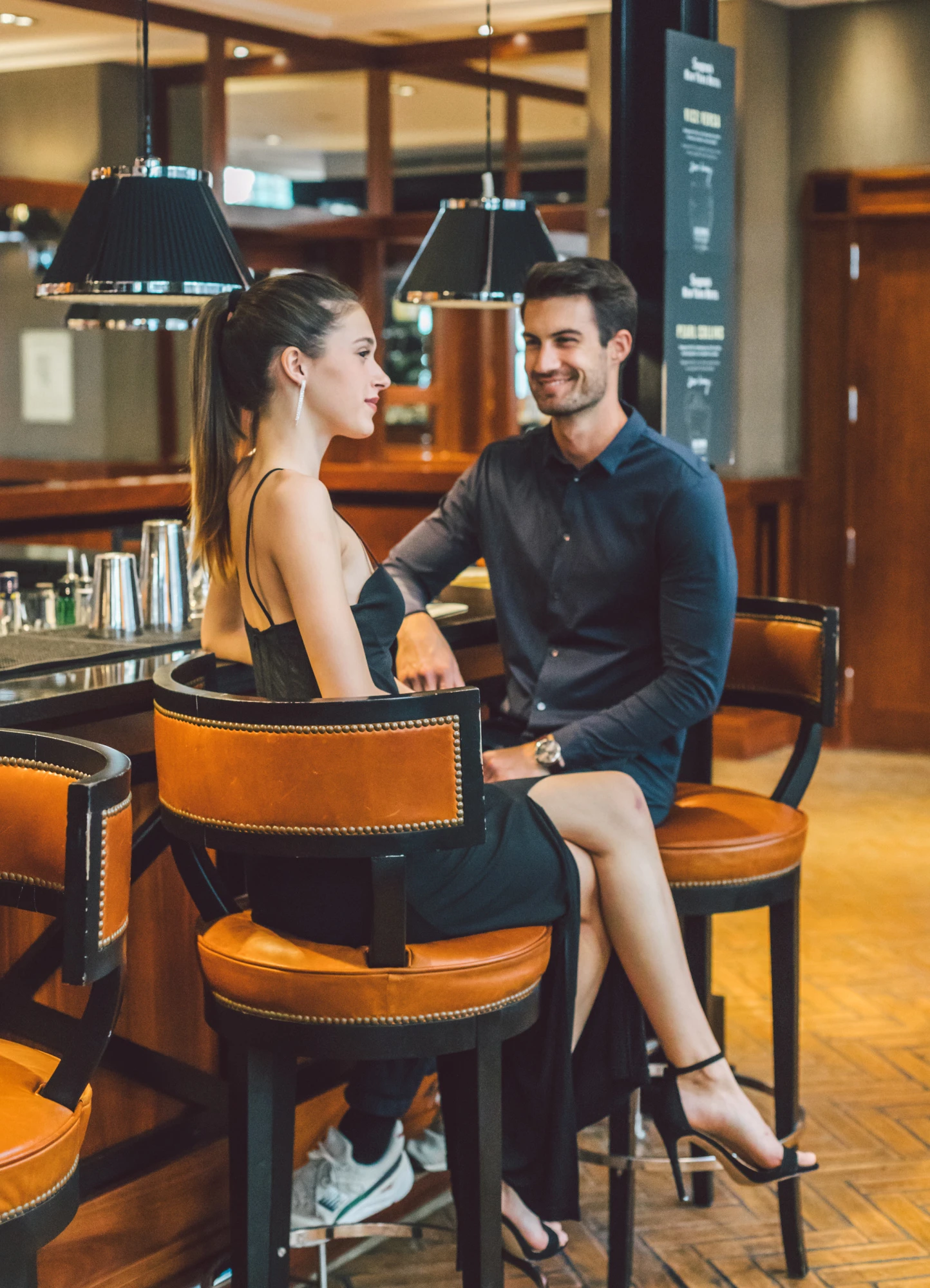 A couple is chatting while seated on chairs in a bar, with the counter and stools in the background.