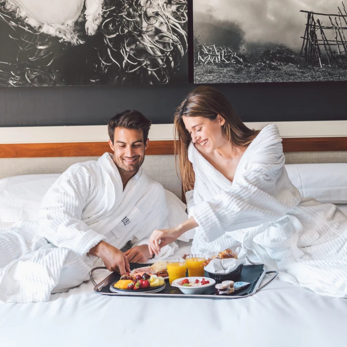 Una pareja desayuna en una bandeja sobre una cama con sábanas blancas.