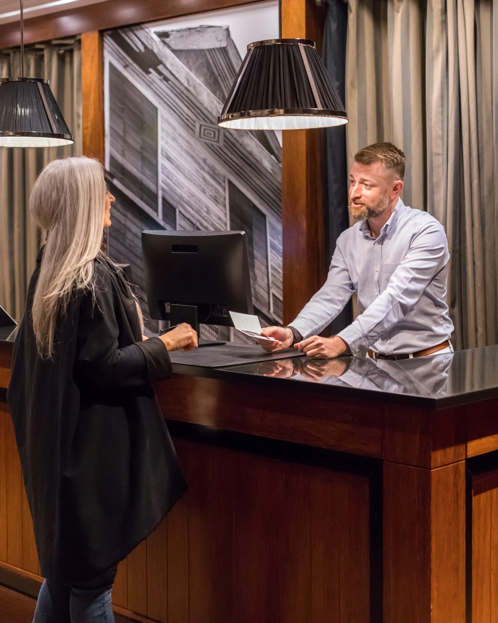 Una recepcionista atiende a un huésped en un mostrador de recepción con lámparas de pie y cortinas de fondo.