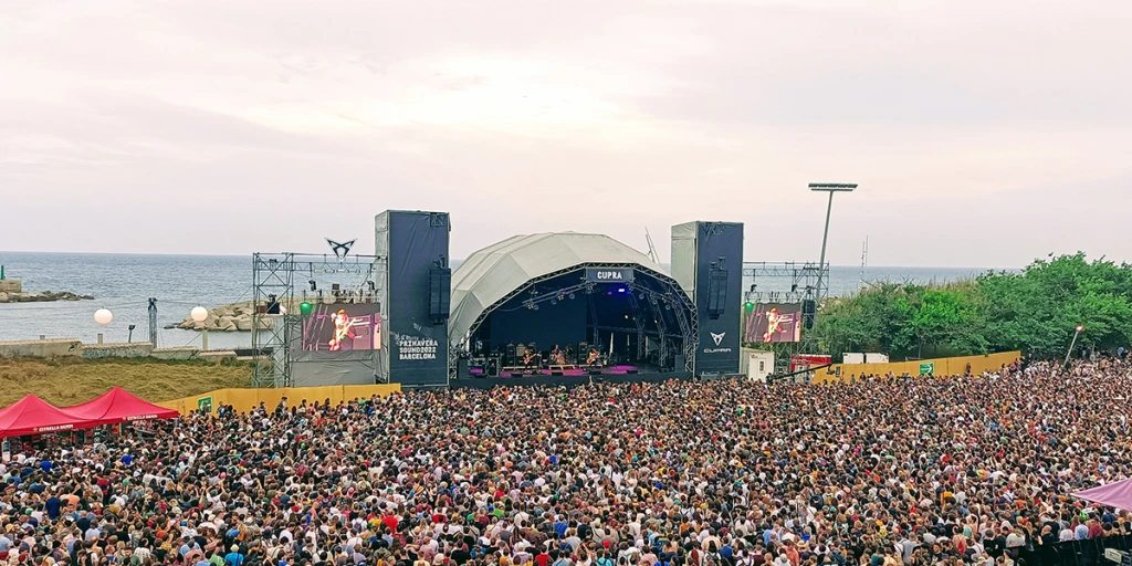 Una multitud asiste a un festival al aire libre frente al mar con un escenario iluminado.