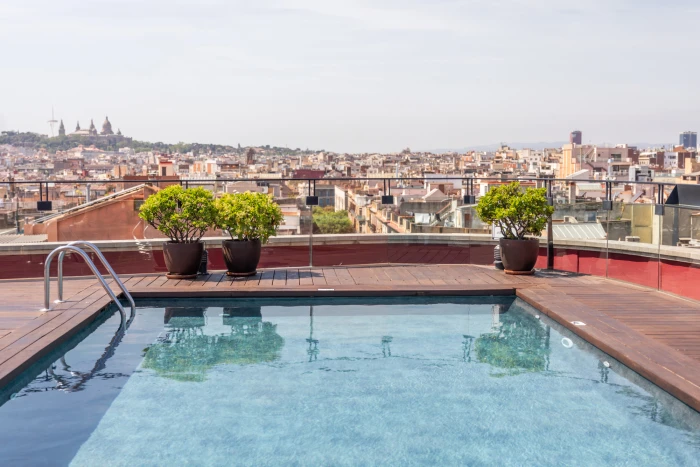 On voit une piscine sur une terrasse avec des garde-corps et des plantes en pot, avec la ville en arrière-plan.