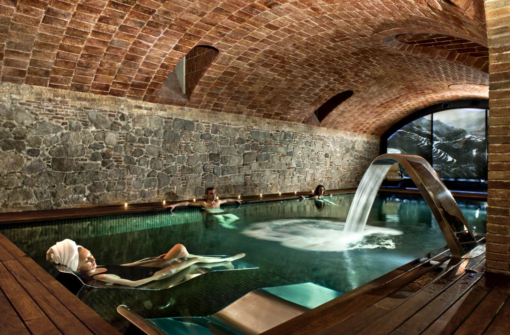 Piscina cubierta con agua verde y chorros, rodeada por paredes de piedra y suelo de madera.
