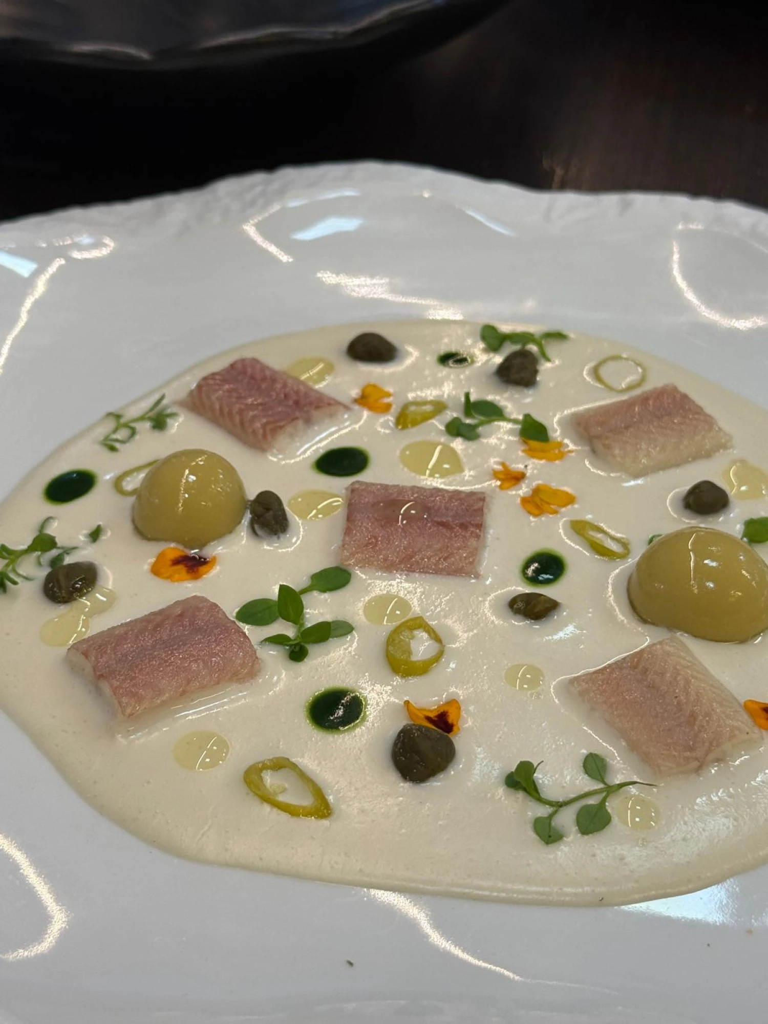 Una presentación elegante de un plato de ajoblanco de coco, decorado con esferas de aceituna y anguila ahumada, resaltando su estilo sofisticado y moderno.