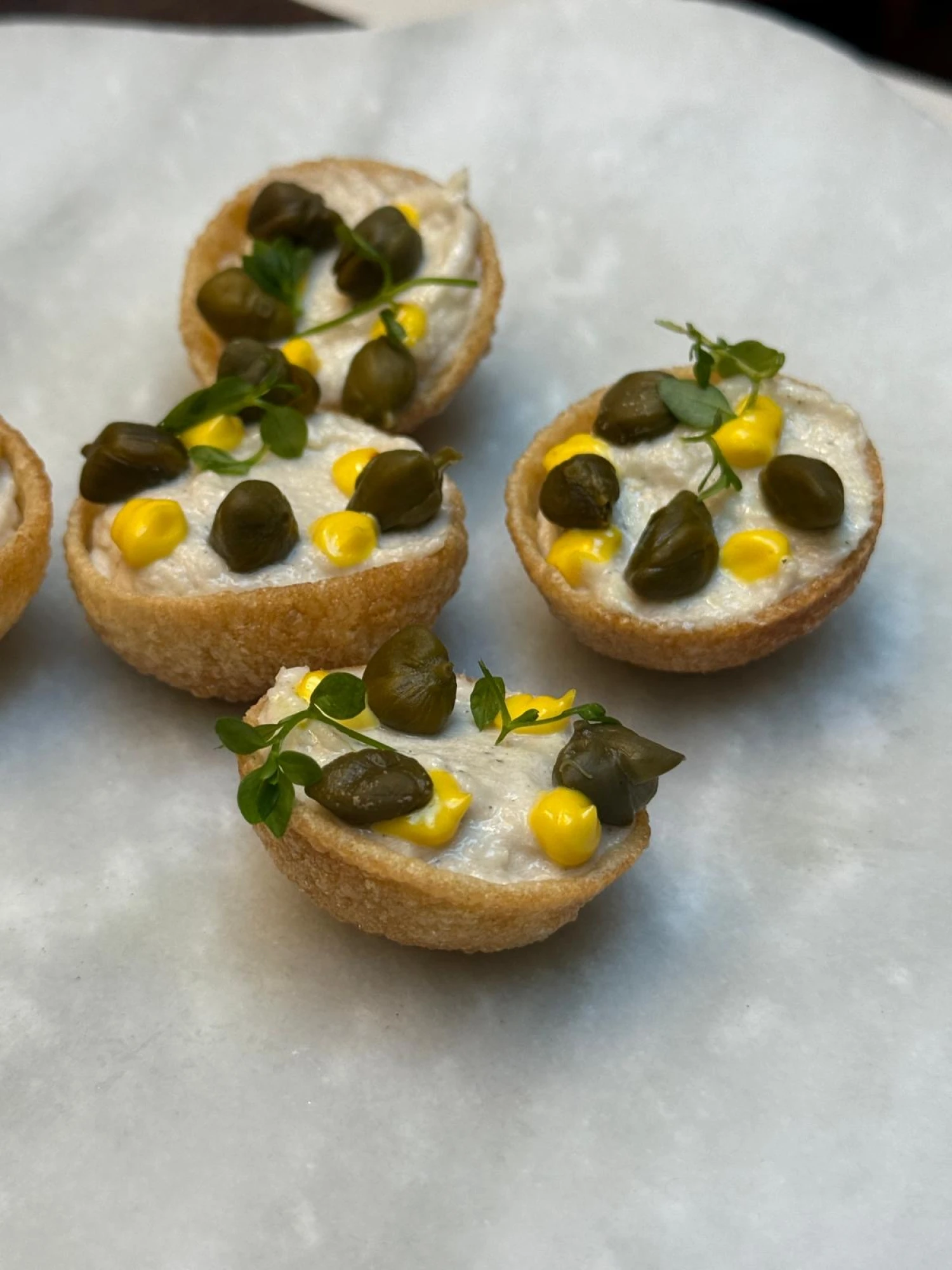 Una presentación de pani puri relleno con una cremosa brandada de bacalao, decorado con alcaparras y acompañado de un alioli de azafrán, ideal para un aperitivo sofisticado.