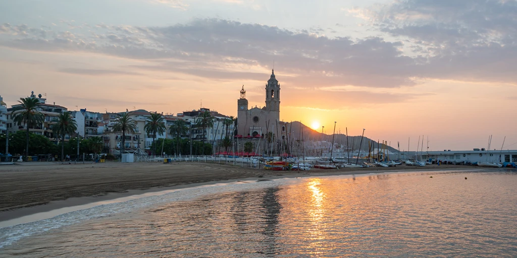 Es veu una platja amb aigua tranquil·la i vaixells amarrats mentre el sol es pon a l'horitzó.