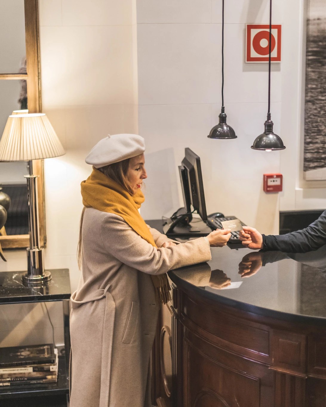 A receptionist assists a guest at a wooden counter with lamps and terminals on the table.