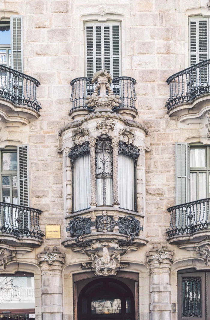 On voit une façade en pierre avec des fenêtres à volets et un balcon en fer forgé orné de décorations sculpturales.