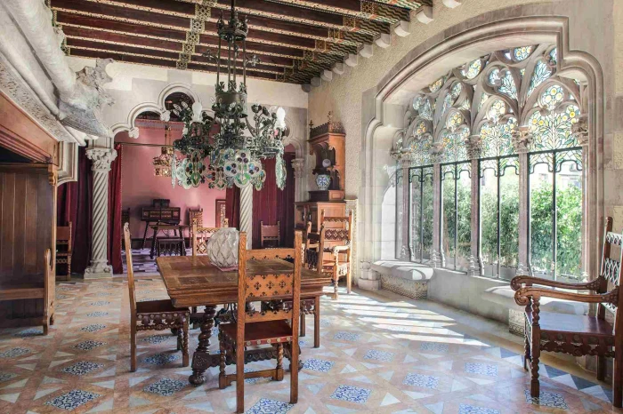 Si vede un ampio salone con tavoli in legno, sedie, pavimento in mosaico e grandi finestre ad arco illuminate dalla luce naturale.