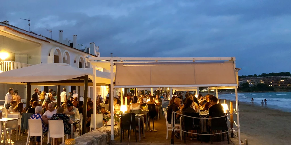 Zu sehen ist eine Terrasse mit Tischen und Stühlen unter Markisen, direkt am Meeresufer bei Sonnenuntergang.