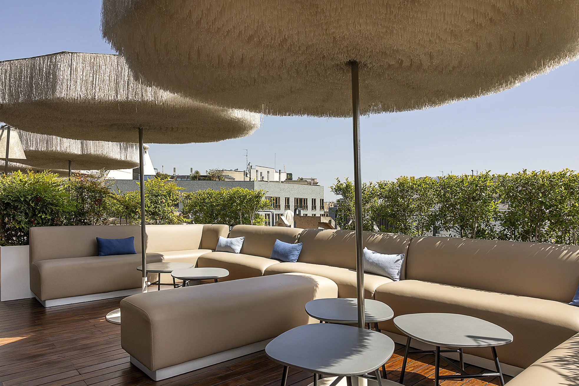 Se ve una terraza con sofás y mesas bajo sombrillas, junto a setos y vista a edificios al fondo.