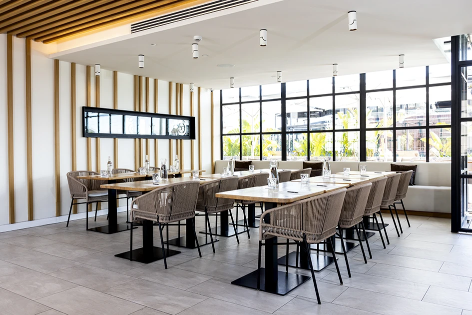 Se ve un comedor con mesas, sillas y grandes ventanales con luz natural.
