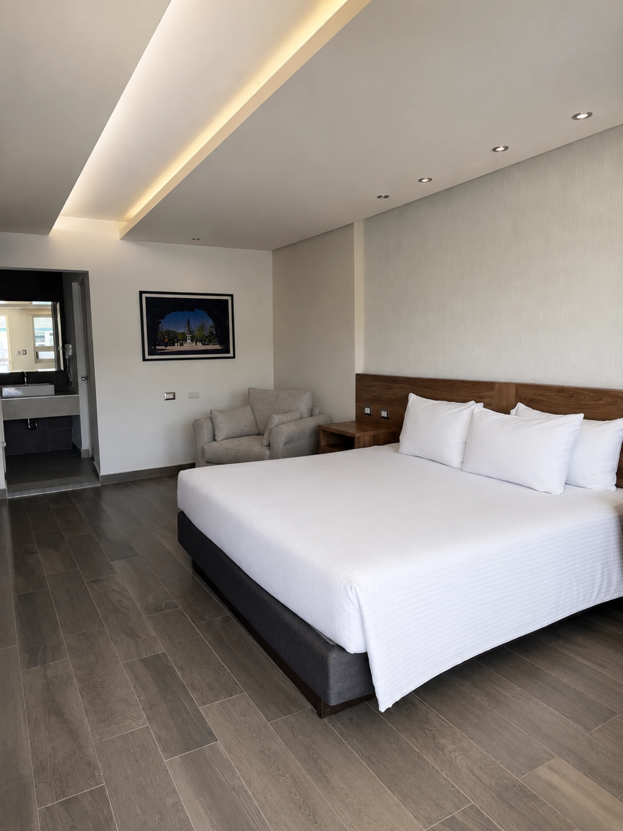 Se ve una cama doble con ropa de cama blanca y un sofá gris en un dormitorio con suelo de madera oscura.
