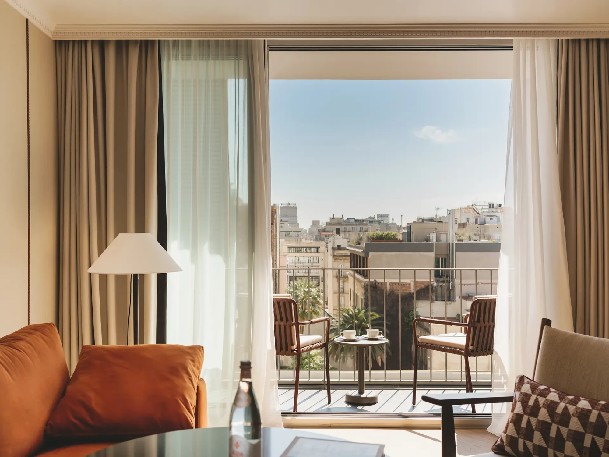 Se ve una sala con sofá, mesa y dos sillas junto a puertas de vidrio que dan a un balcón con vista urbana.