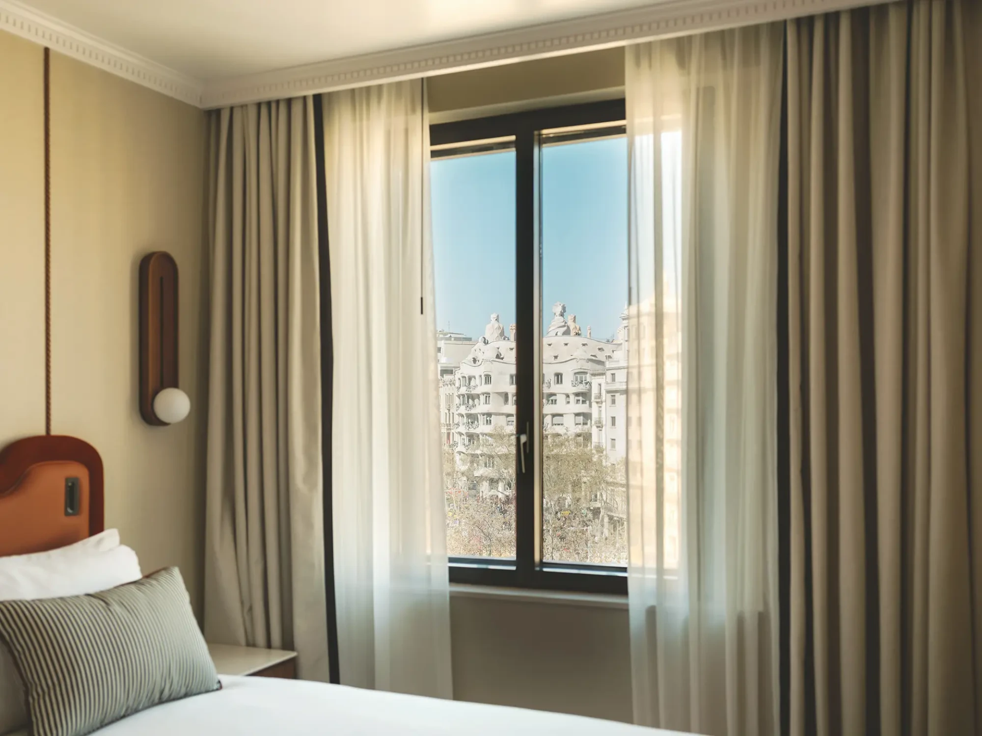 Se ve una cama con almohadas y una ventana con cortinas y luz natural.