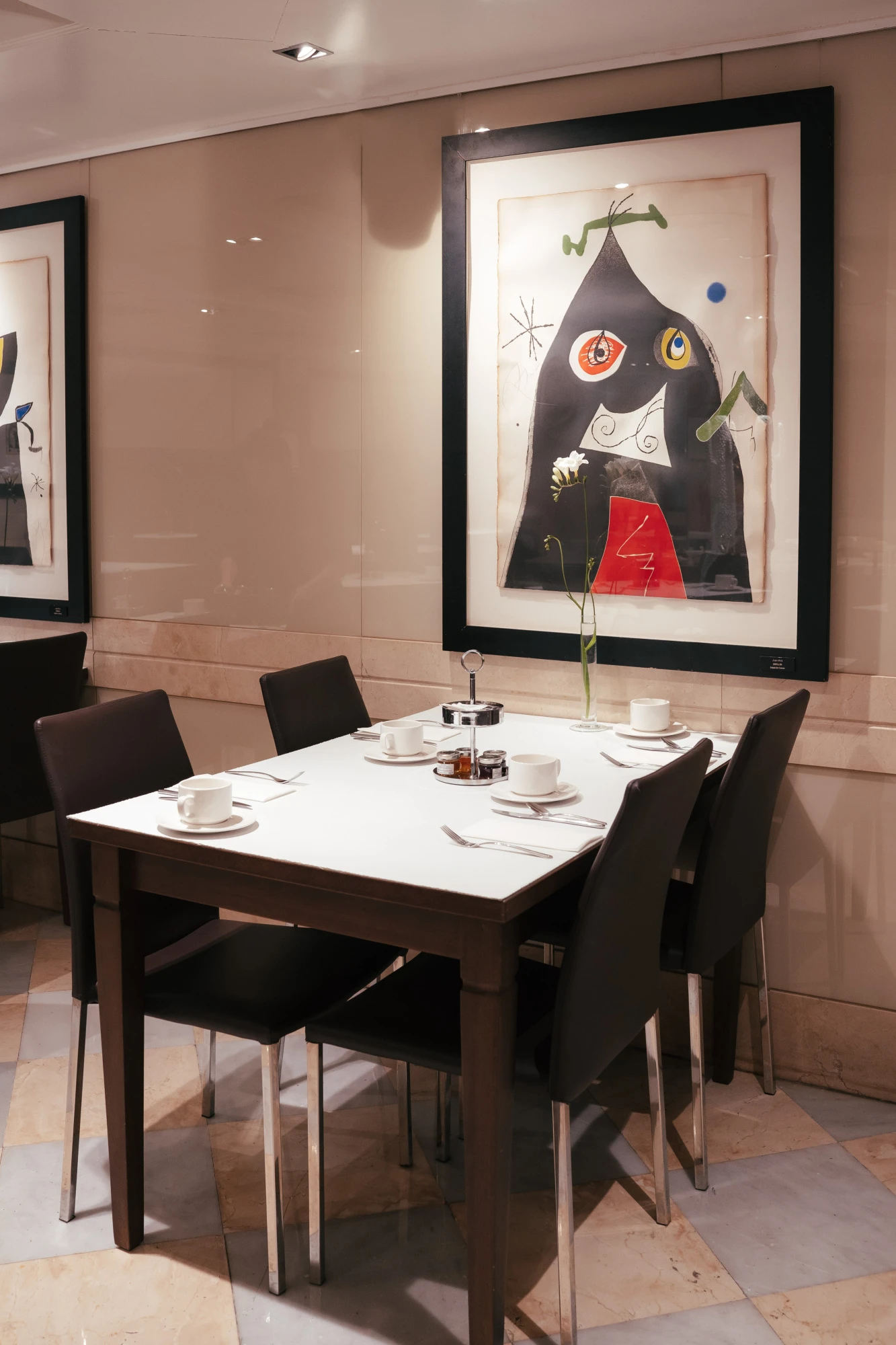 Se ve una mesa de comedor con vajilla y cuatro sillas, junto a cuadros de Joan Miró en una pared clara.
