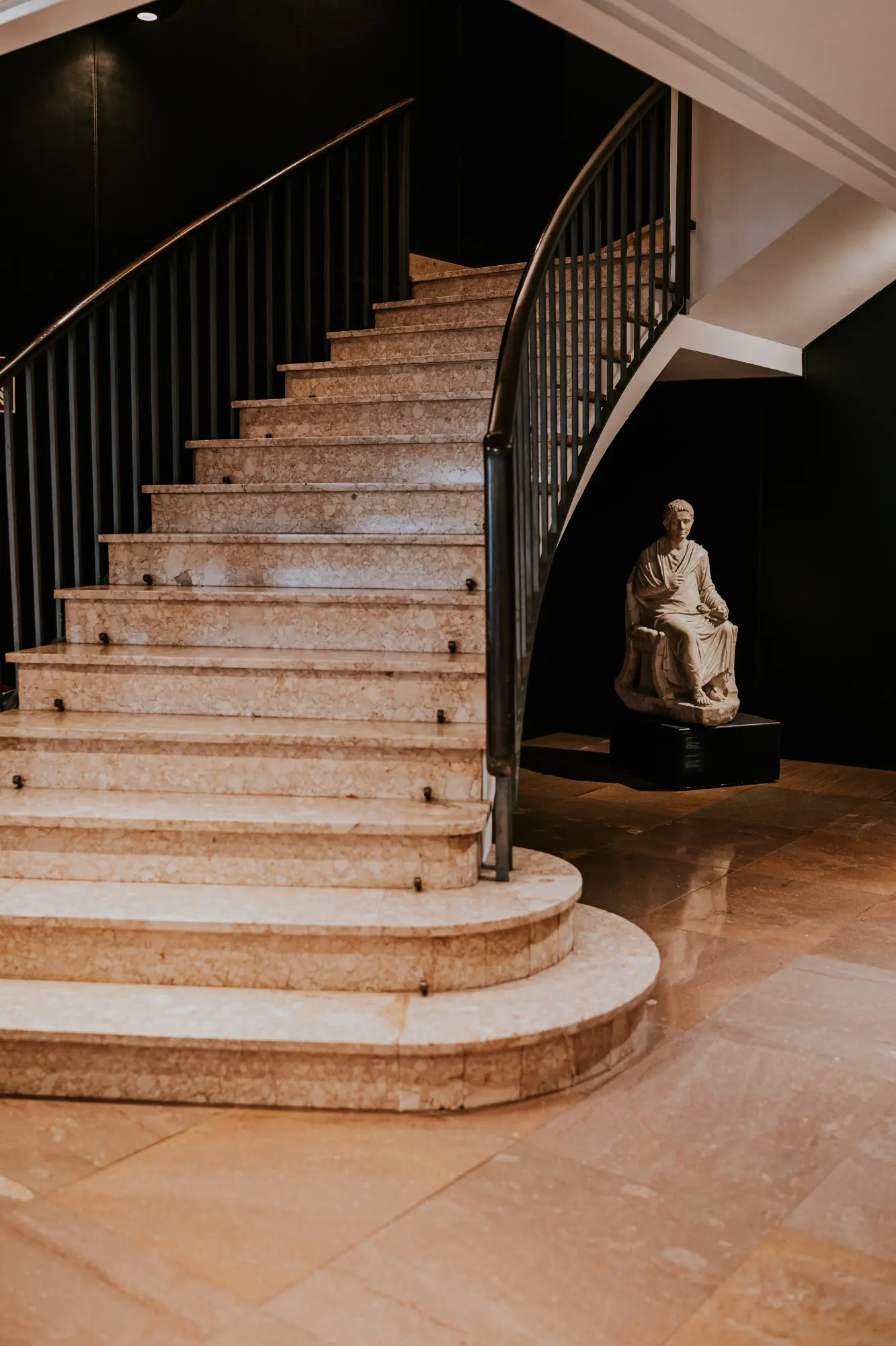 Se ve una escalera de piedra con barandillas negras y una estatua romana sobre un pedestal en el vestíbulo.