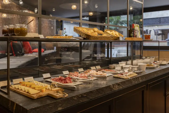 Se ve una mesa de buffet con bandejas de comida y utensilios en un comedor con iluminación de techo.