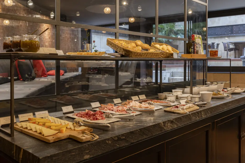 Se ve una mesa de buffet con bandejas de comida y utensilios en un comedor con iluminación de techo.