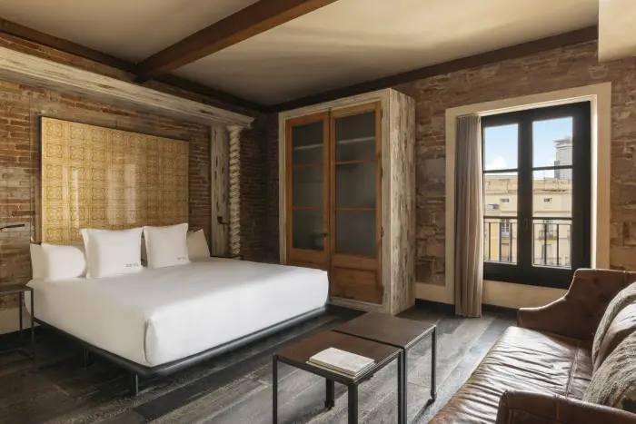 Se ve una cama doble con ropa de cama blanca, un sillón marrón y una ventana con luz natural en la habitación.