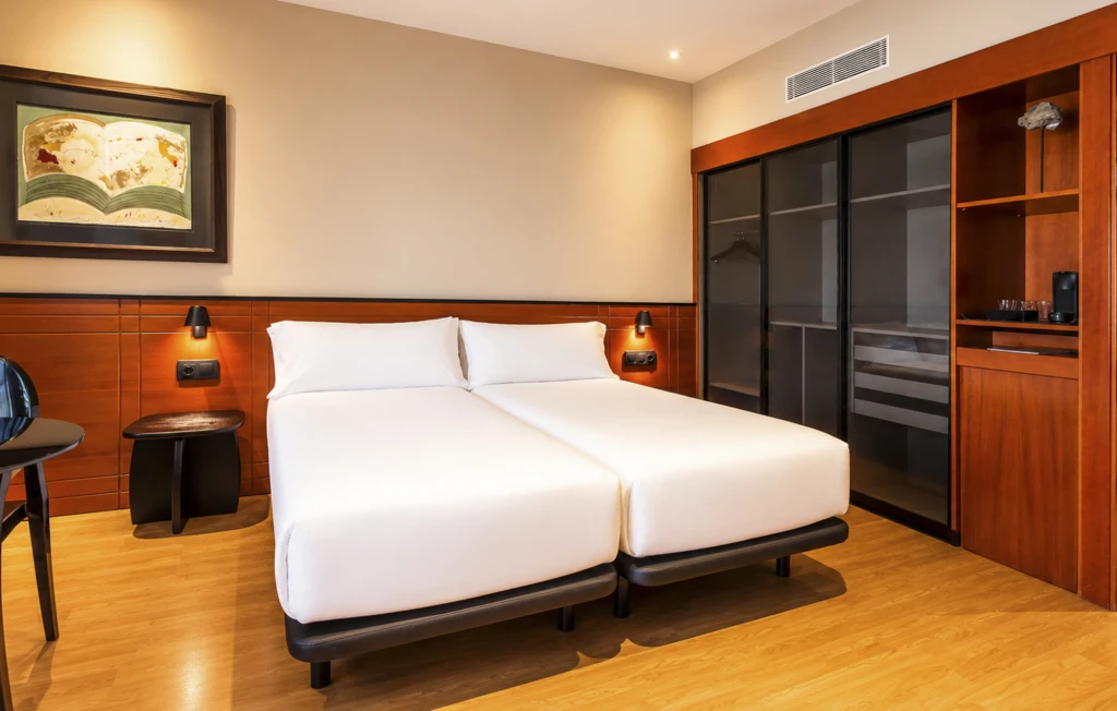 Se ve una cama doble con ropa de cama blanca, mesita y lámparas junto a una pared de madera y un armario empotrado.