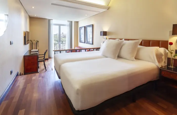 Se ve una cama doble con ropa de cama blanca y un ventanal con luz natural en una habitación moderna.