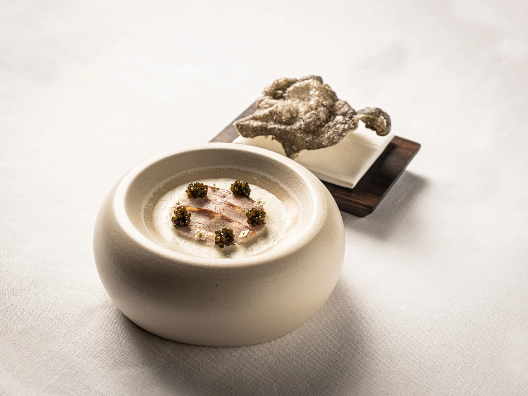 Se ve un plato blanco con una crema y pequeñas porciones de caviar sobre una mesa clara.
