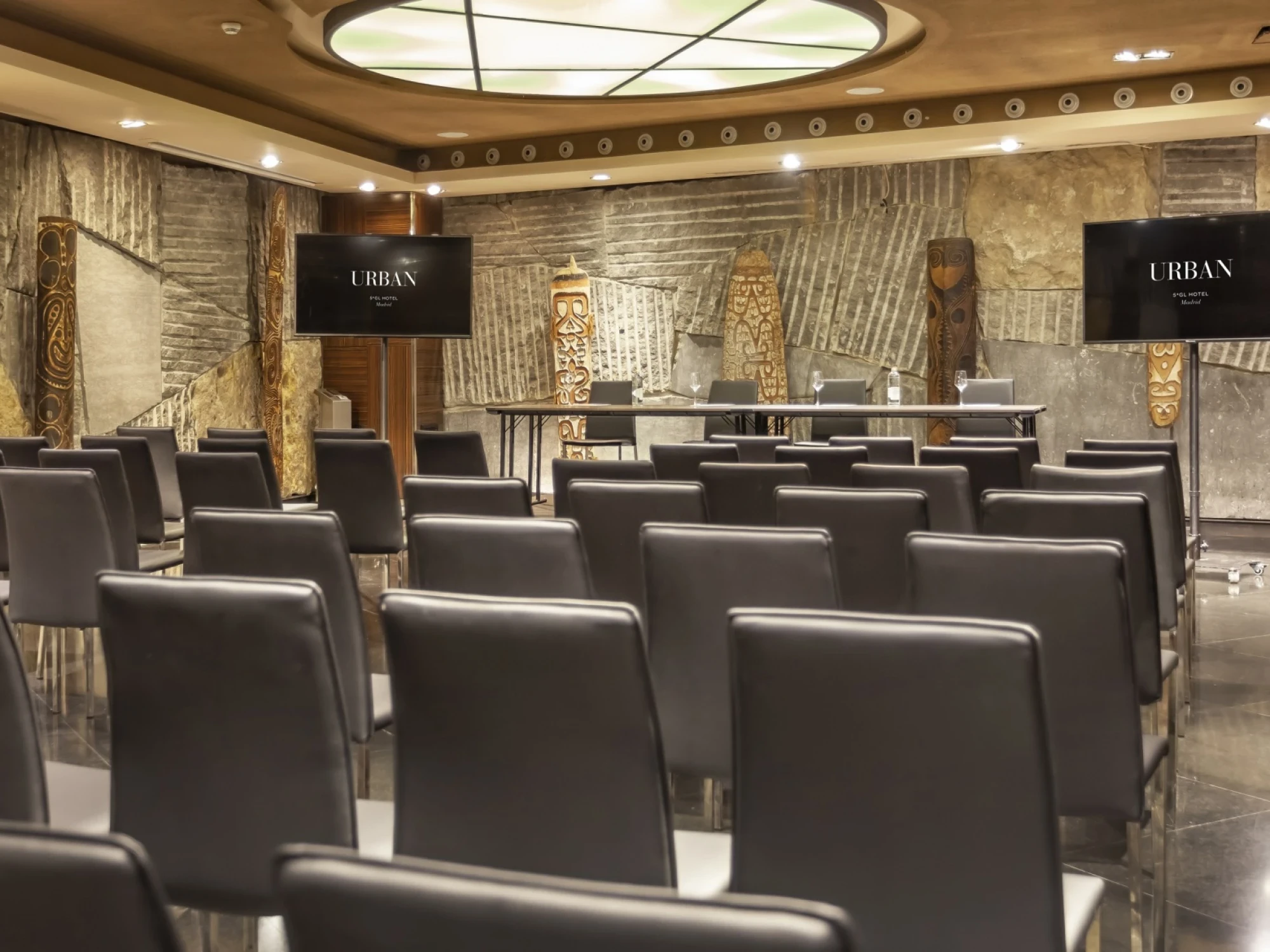 Se ve una sala de reuniones con filas de butacas grises y un escenario al fondo con pantallas.