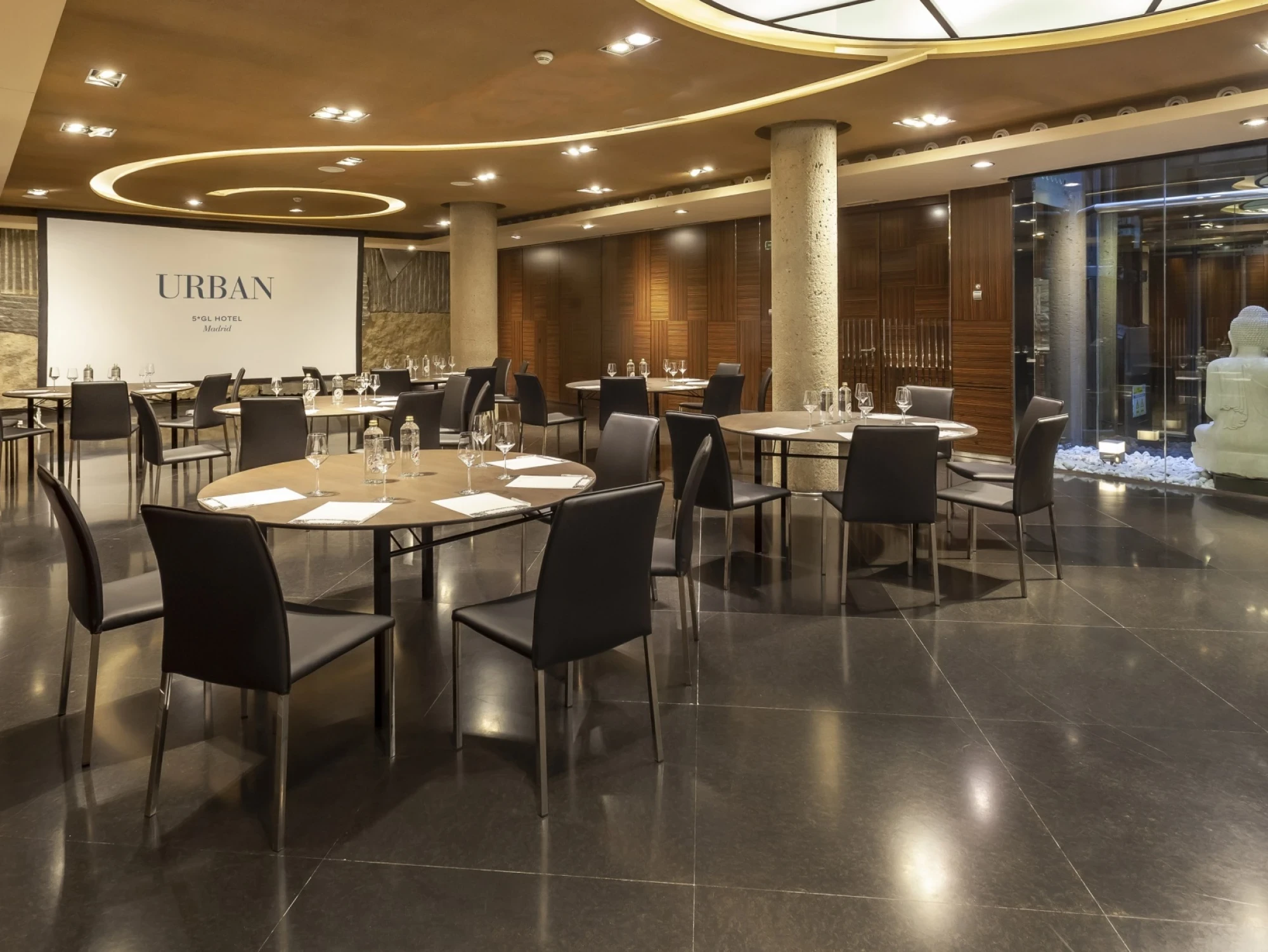 Se ve un comedor interior con mesas con manteles claros y sillas negras alrededor, con iluminación empotrada en el techo.
