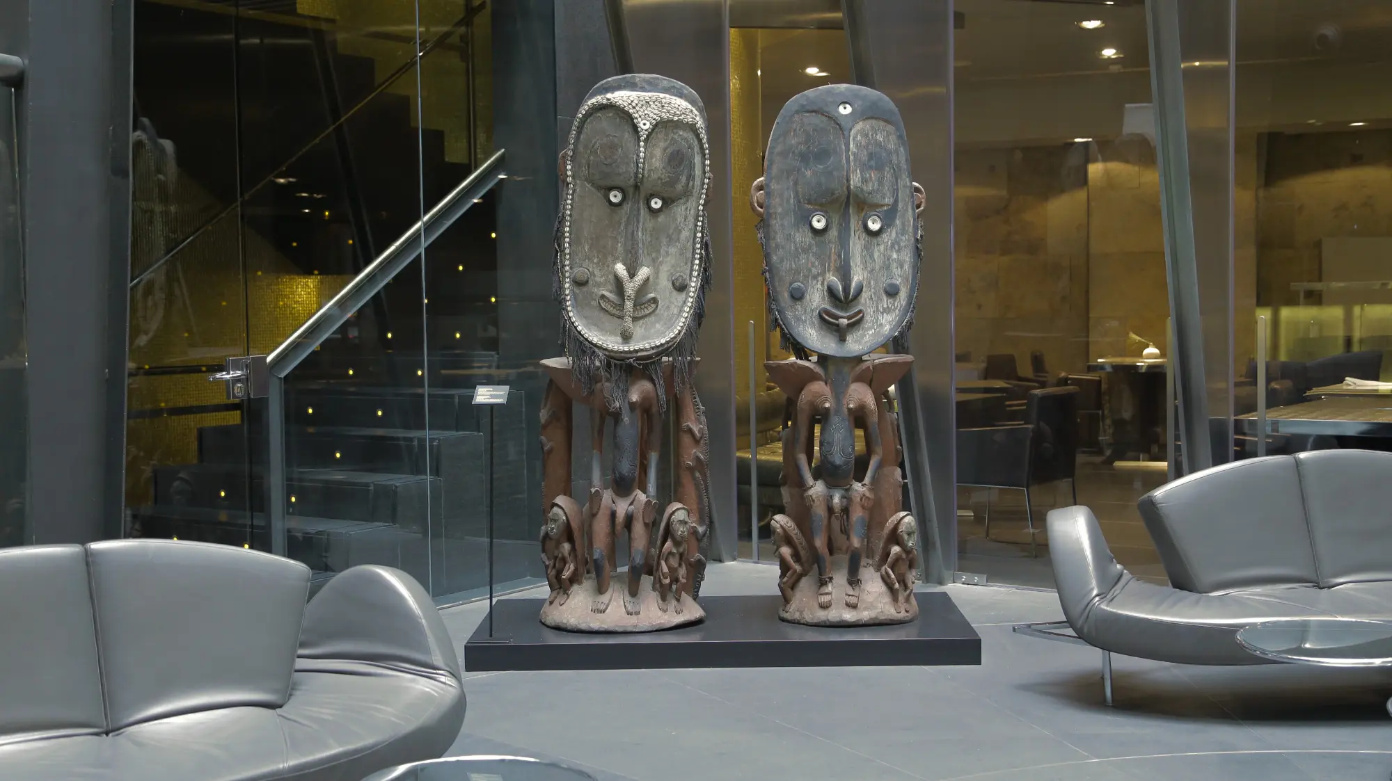Se ve una zona de estar con sillones grises y esculturas decorativas sobre pedestales junto a un ventanal.