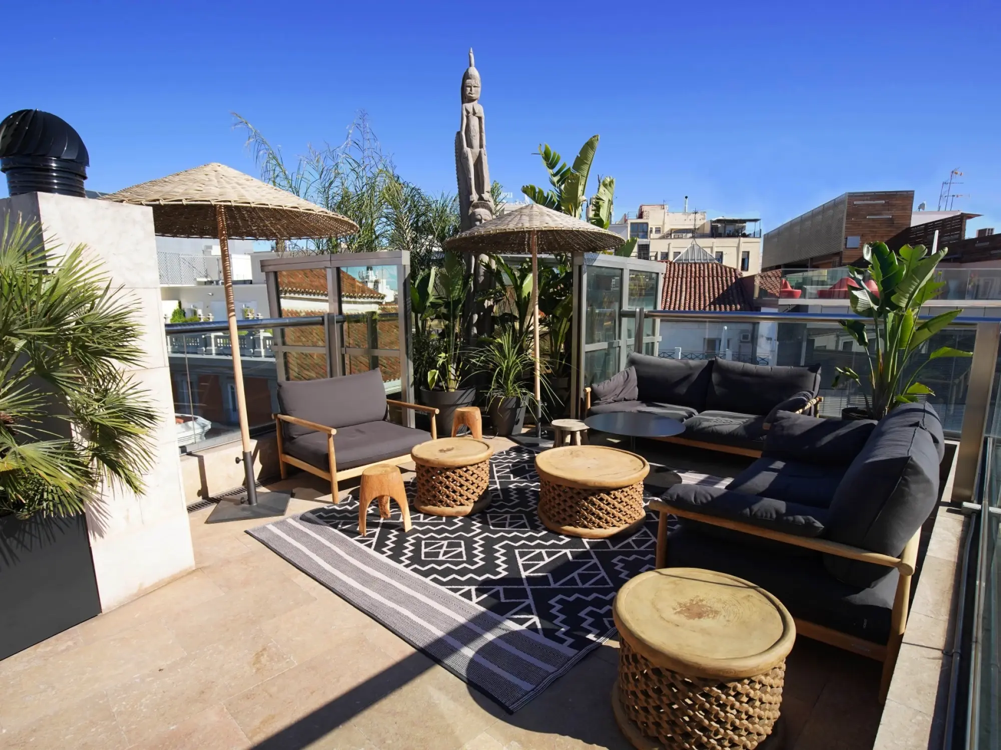 Se ve una terraza al aire libre con tumbonas, mesas auxiliares y sombrillas sobre un suelo de madera y alfombra decorativa.