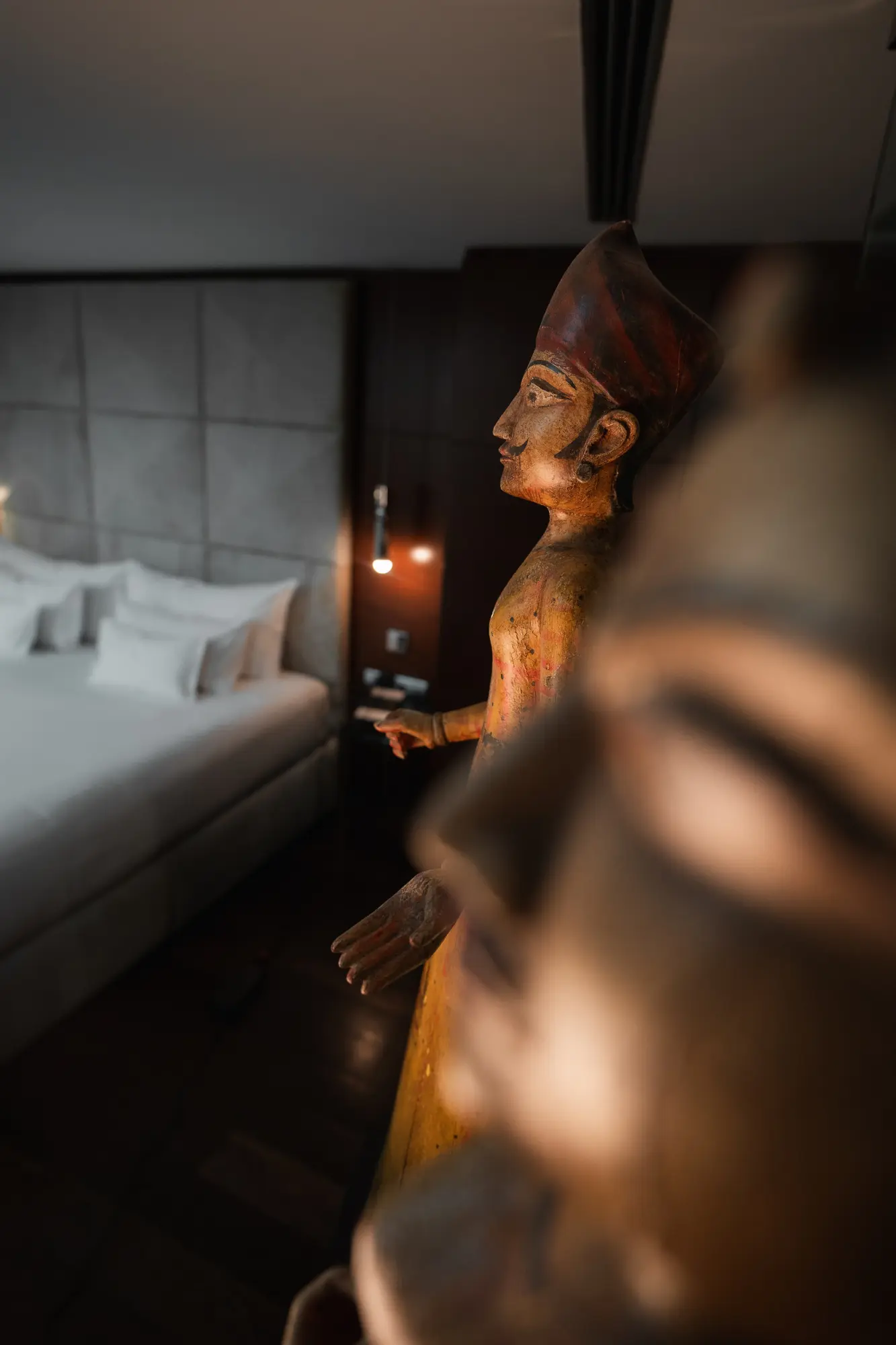 Se ve una escultura decorativa de madera junto a una cama en una habitación con iluminación tenue.