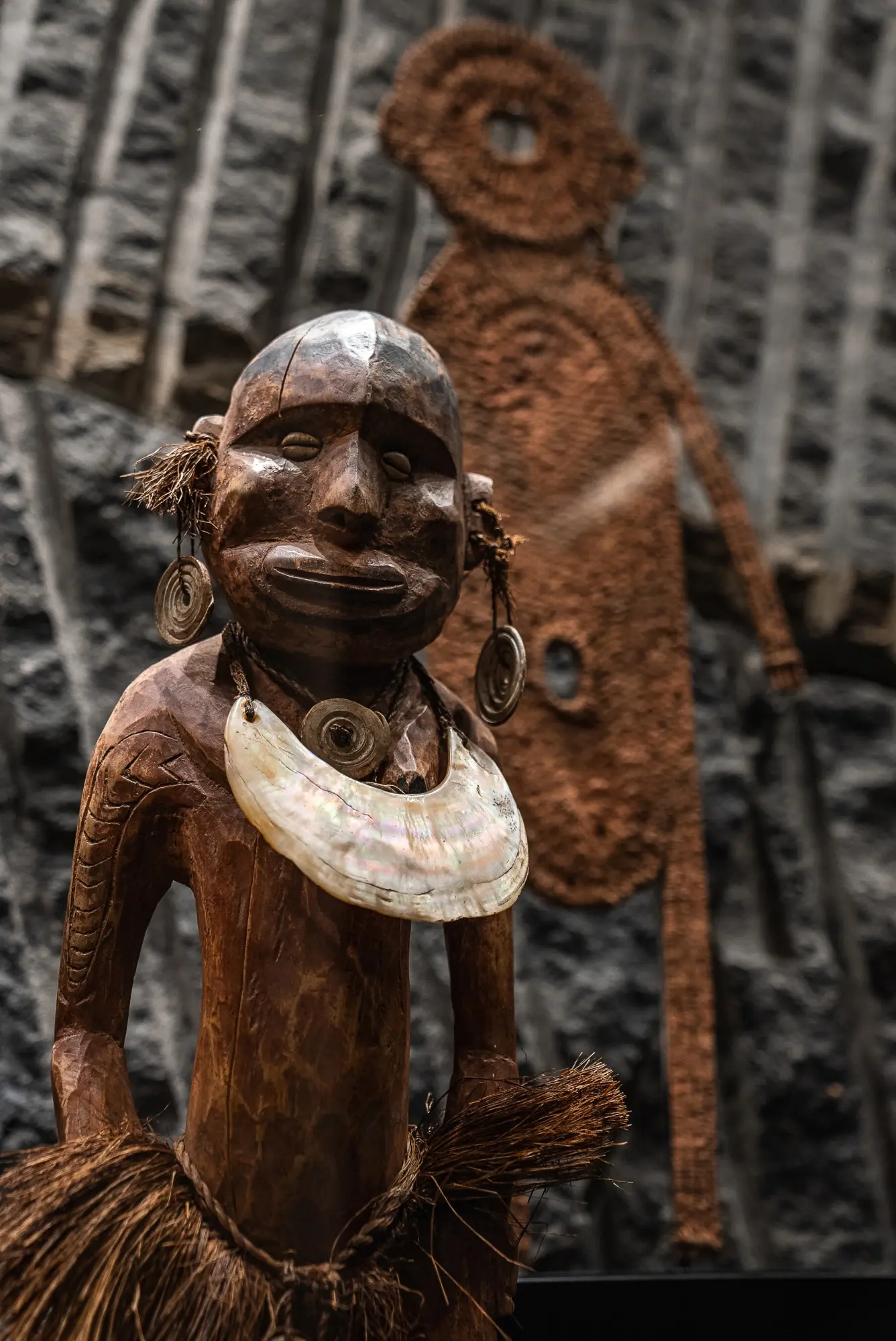 Se ve una escultura decorativa de madera con un tocado y un collar, sobre un fondo con figuras talladas.