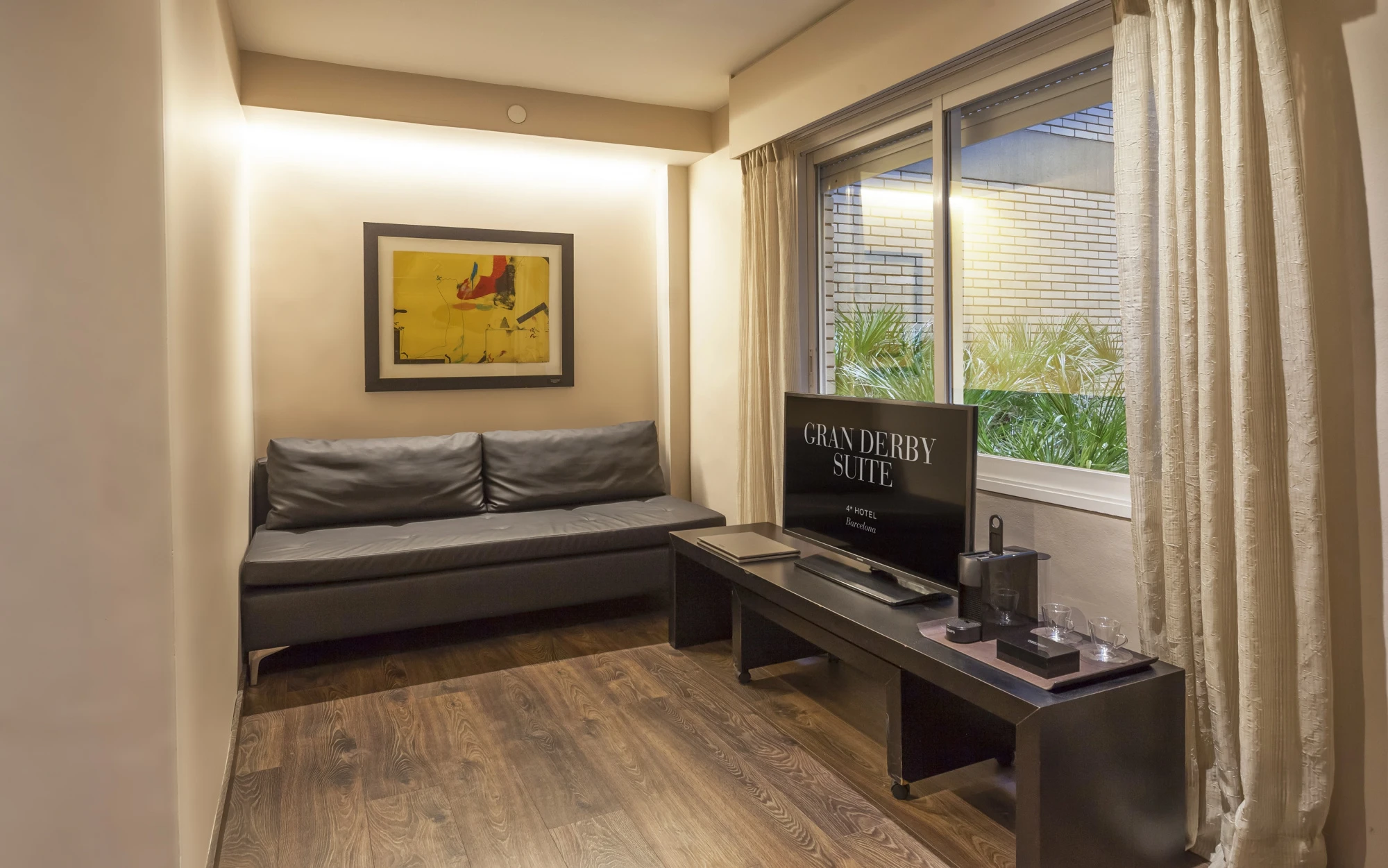 Se ve una cama con cojines, una mesa auxiliar y una ventana con cortinas y planta en el alféizar.