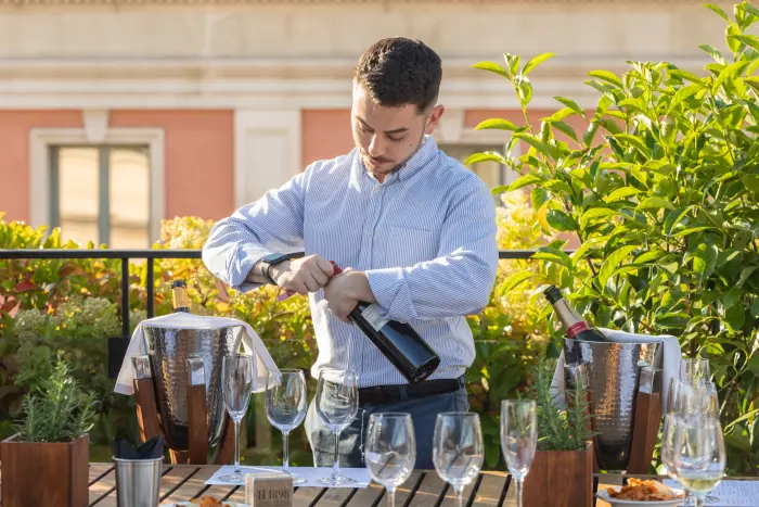 A waiter pours drinks from a bottle at an outdoor table with glasses and carafes.
