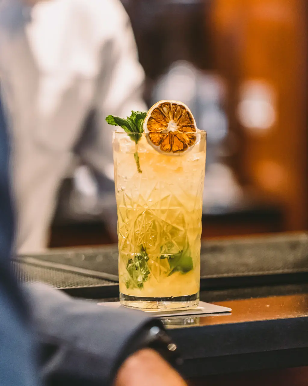 They serve a cocktail with an orange slice and herbs on the bar counter.
