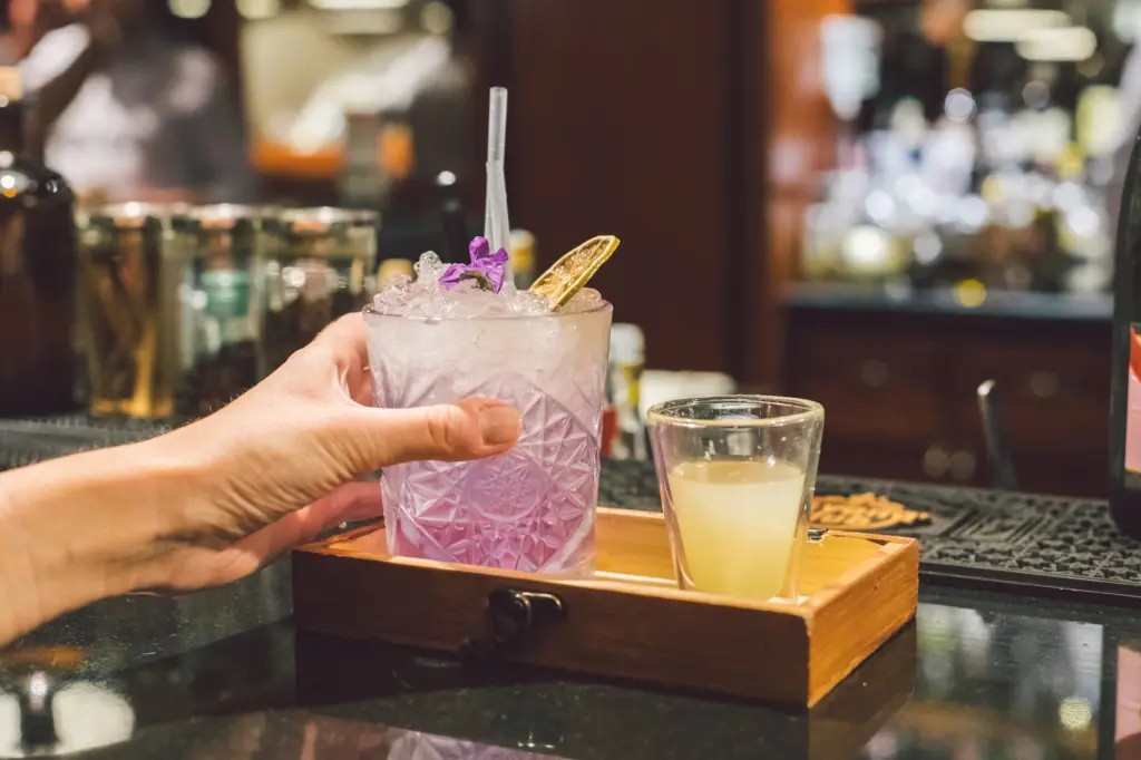 A hand holds a glass with ice and a straw over a counter, beside a drink in a glass.