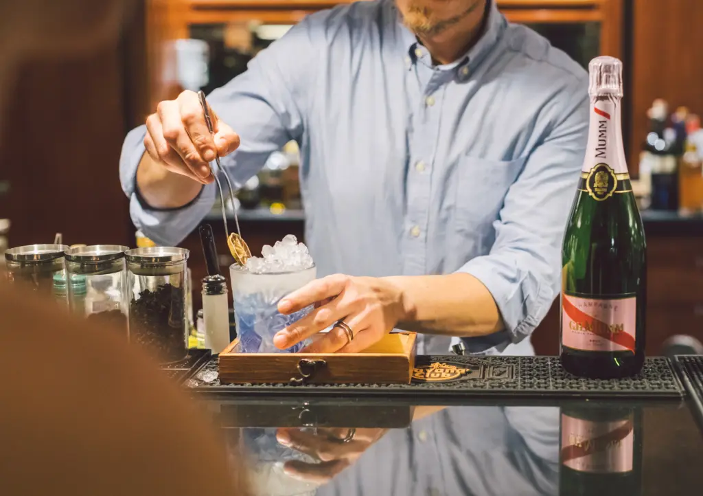 A waiter prepares a drink beside bottles and glasses on the bar counter.