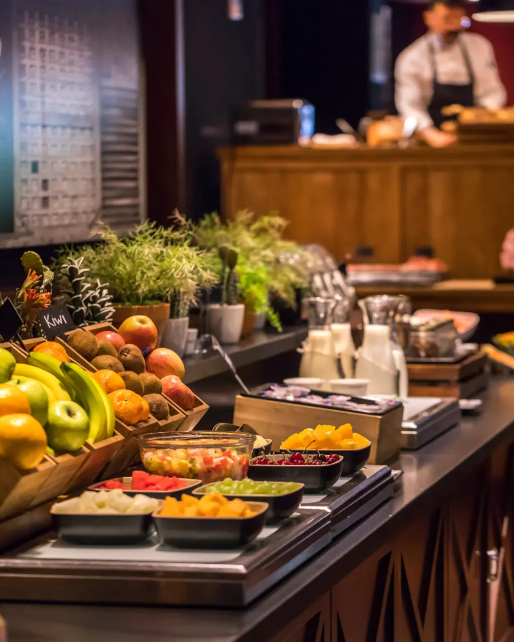 Una varietà di alimenti in un buffet per la colazione, tra cui frutta, pane e altri prodotti, presentati in un ambiente luminoso e accogliente.