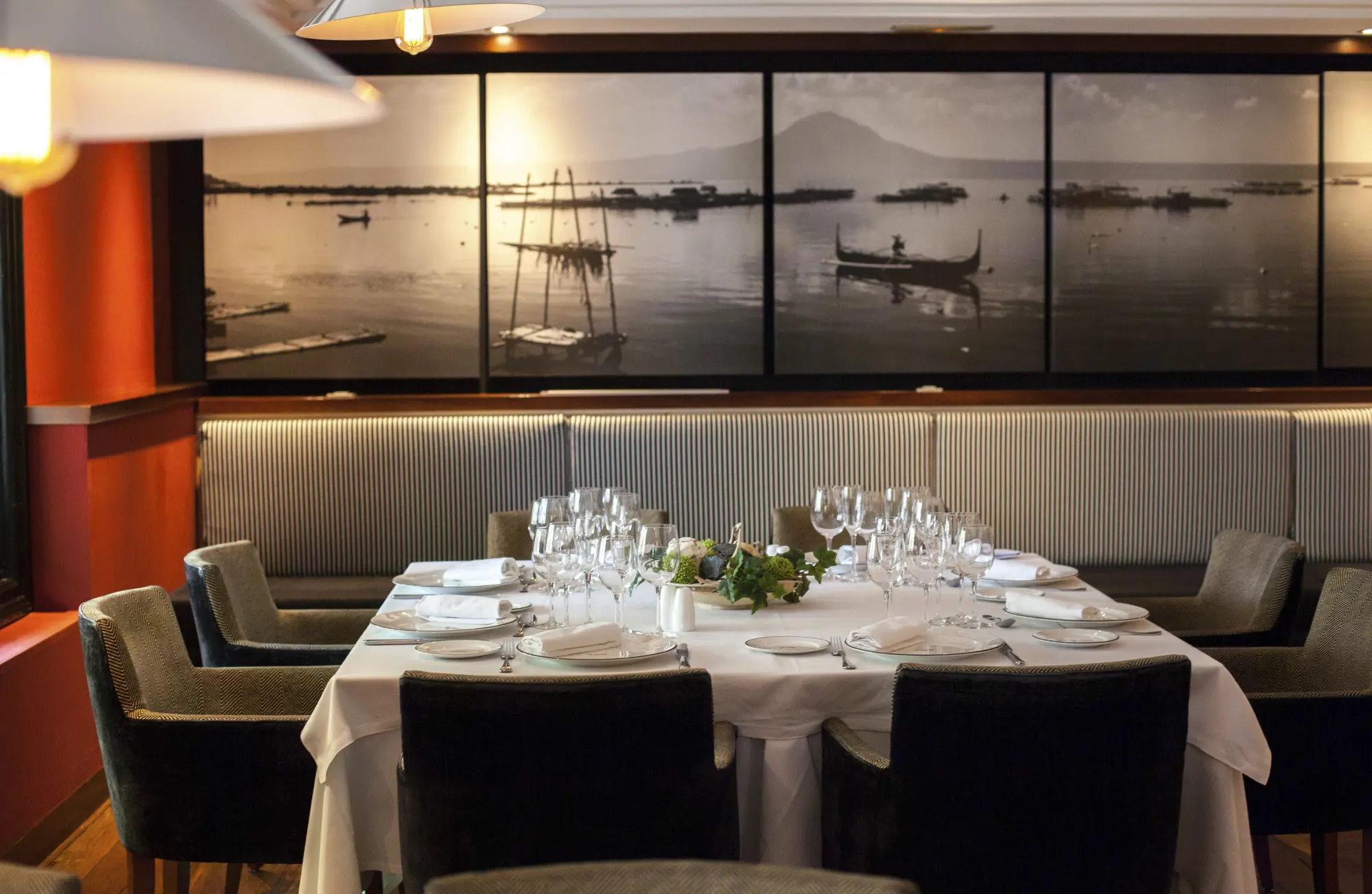 Una mesa puesta con copas y platos se ve en un comedor con asientos tapizados y decoración marina en la pared.