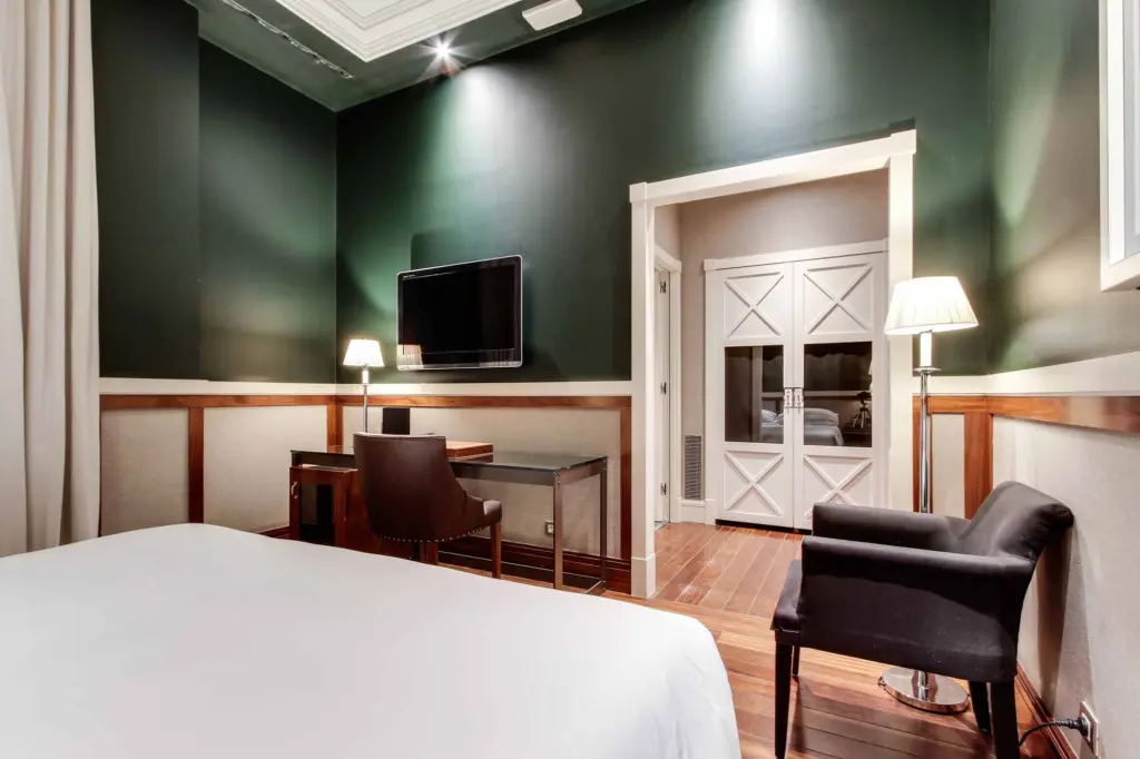 Se ve una cama, un escritorio con silla y una televisión en una habitación con paredes verdes y lámparas de pie.