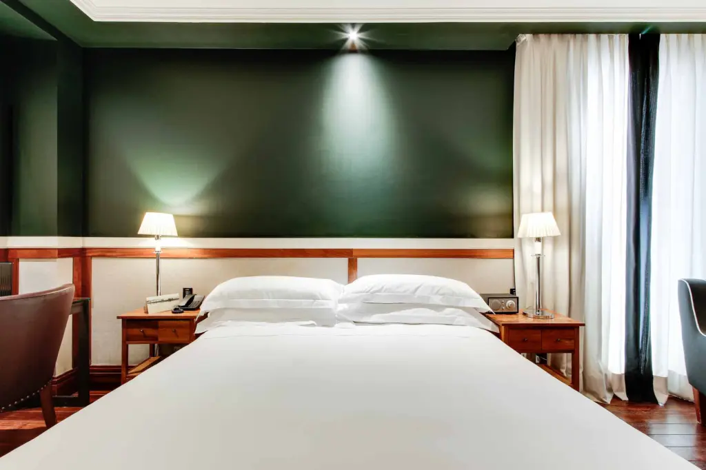 Es veu un llit doble amb tauletes de nit i làmpades, juntament amb cortines i una paret verda a l'habitació.