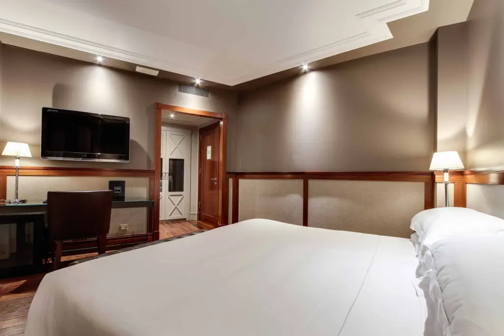 Es veu una habitació amb un llit doble, dues tauletes de nit i làmpades, i un televisor a la paret.