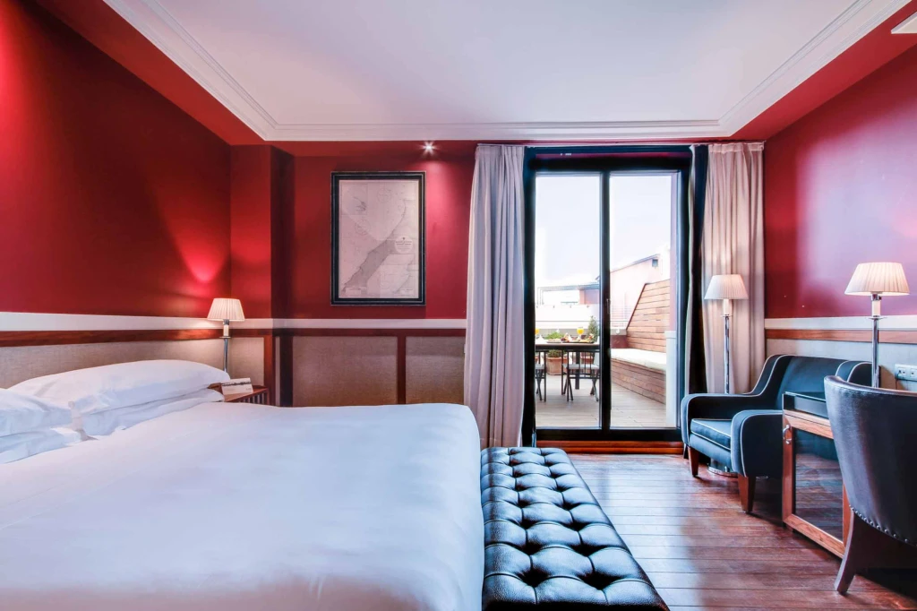 The image shows a double bed, bedside tables with lamps, an upholstered bench, and a door leading to the balcony.
